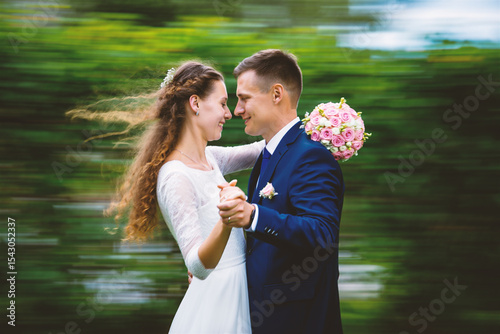 A romantic wedding couple dances outdoors with a blurred, dynamic green background. They hold hands and look at each other, celebrating their love and happiness on their special day.