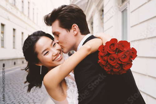 Happy wedding couple embracing on a street, man kisses woman's cheek, she holds red rose bouquet. Love and romance outdoors.