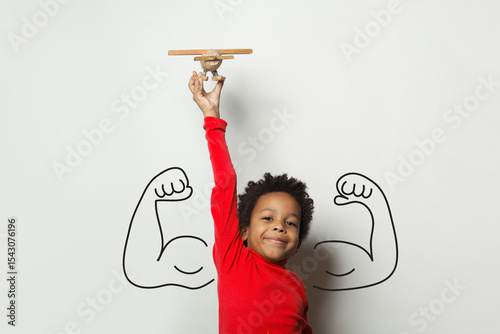 Creative Child Plays With Wooden Airplane and Poses Confidently in Front of a Strong Muscle Mural