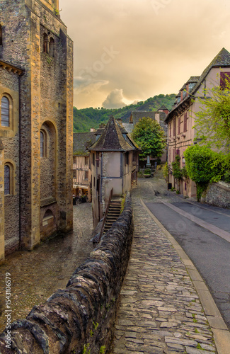 Fototapeta Naklejka Na Ścianę i Meble -  Ancient medieval cobblestone streets with a farmhouse on the corner and the Abbey of Sainte-Foy in the historic village of Conques, with mountains in the background under a spectacular sunset sky.
