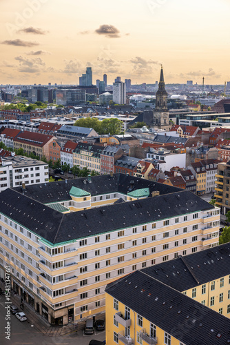 Panoramic view of Copenhagen taken from the top of the Church of Our Saviour, showing a mix of historical rooftops, modern buildings, and city landmarks under a warm evening sky.