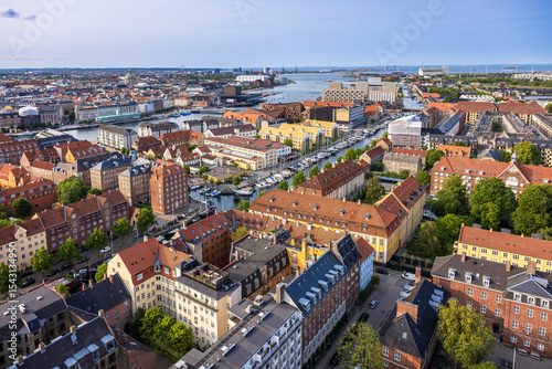Panoramic view of Copenhagen taken from the top of the Church of Our Saviour, showing a mix of historical rooftops, modern buildings, and city landmarks under a warm evening sky.