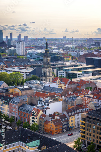 Panoramic view of Copenhagen taken from the top of the Church of Our Saviour, showing a mix of historical rooftops, modern buildings, and city landmarks under a warm evening sky.