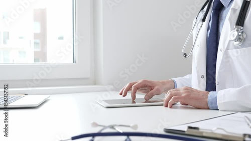 Male doctor wearing lab coat and stethoscope using digital tablet while sitting at desk in medical office, with medical chart and pen in the foreground