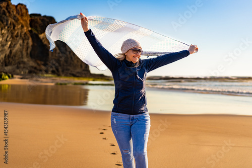 Beautiful middle aged woman strolling holding shawl on sandy beach on spring afternoon. Monte Clerigo Praia, Portugal. Front view