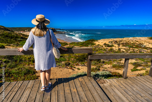 Mature woman in summer hat tourist standing at viewpoint to Monte Clerigo beach on Algarve coast on sunny spring day. Back view	
