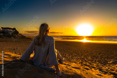 Beautiful middle-aged woman sitting on sandy beach on sunset on spring day. Back view. Monte Clerigo beach on Algarve coast in Portugal