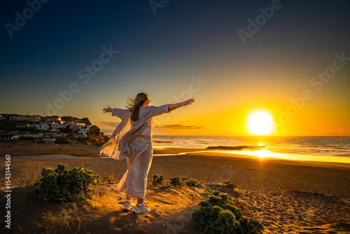 Beautiful middle-aged woman standing with outstretched arms on sandy beach on sunset on windy spring day. Back view. Monte Clerigo beach on Algarve coast in Portugal