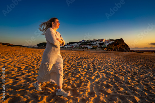 Beautiful middle-aged woman walking on sandy beach on sunset on windy spring day. Side view. Monte Clerigo beach on Algarve coast in Portugal