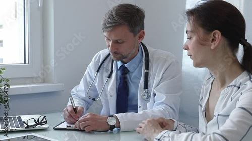 Male doctor writing notes on a clipboard while attentively listening to a female patient during a medical consultation in a hospital office, fostering communication and understanding
