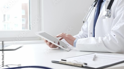 Professional doctor man wearing medical coat and stethoscope using digital tablet while sitting at desk in medical office, with medical chart and pen in the foreground. Medicine and health care