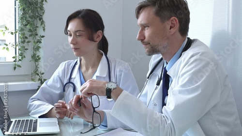 Two doctors are examining medical records on a laptop, engaging in a focused discussion about patient data within a hospital or clinical setting. Medicine and health care concept.