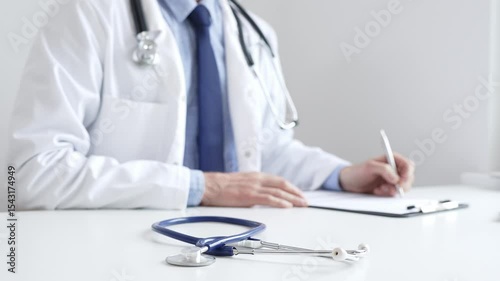 Closeup of blue stethoscope is lying on the table near doctor man professional writing on the clipboard in clinic office. Medicine and healthcare
