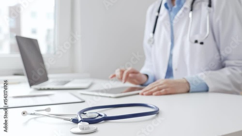 Female doctor wearing lab coat and stethoscope using digital tablet with laptop and medical records on desk in medical office, showcasing modern healthcare technology. Medicine concept