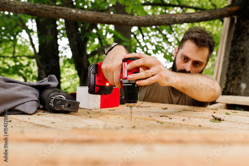 young brunette man, dad with beard is nailing building a tree house to his little son with wooden planks, electric red professional drill, self-tapping screws in the forest