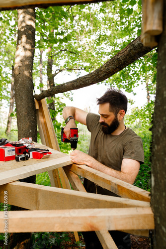 young brunette man, dad with beard is nailing building a tree house to his little son with wooden planks, electric red professional drill, self-tapping screws in the forest