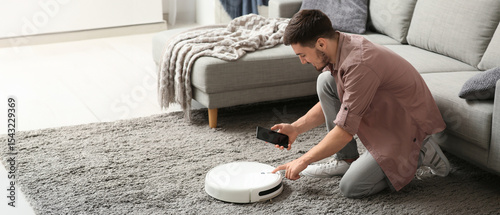 Young man with mobile phone turning on modern robot vacuum cleaner at home