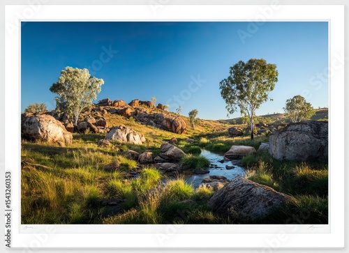 A beautiful landscape of the late afternoon in Australia's outback, with a clear blue sky, boulders and rocks on the ground surrounded by green vegetation around a small river, and a few eucalyptus 