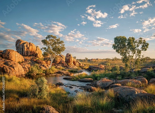 A beautiful landscape of the late afternoon in Australia's outback, with a clear blue sky, boulders and rocks on the ground surrounded by green vegetation around a small river, and a few eucalyptus 