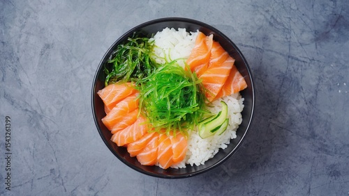 A bowl of sushi rice topped with fresh salmon slices, seaweed, and cucumber. The dish is presented on a gray surface.