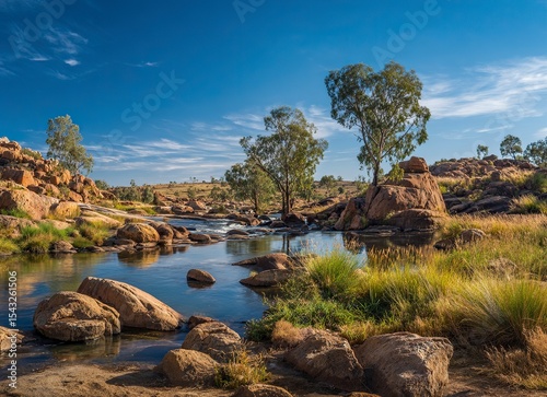 A beautiful landscape of the late afternoon in Australia's outback, with a clear blue sky, boulders and rocks on the ground surrounded by green vegetation around a small river, and a few eucalyptus 