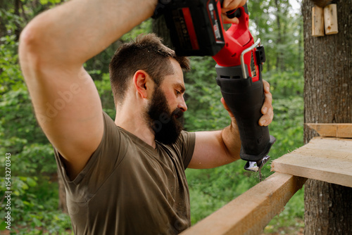 brutal young brunette man, dad with beard is nailing building a tree house to his little son with wooden planks, electric red professional reciprocating saw in the forest. man sawing boards