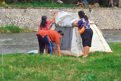 Two mothers and their children work together to properly position the camping tent, while the kids show eagerness to join in photo