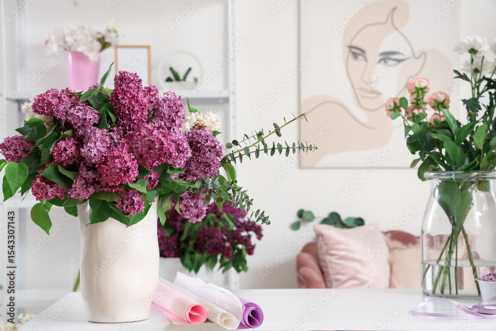 Fototapeta premium Fresh purple lilac flowers in white vase with eucalyptus branches and craft paper rolls on white table, the floral display creates a serene and inviting atmosphere in a modern interior setting
