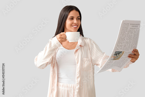 Beautiful woman in pajamas with coffee cup reading newspaper on light background