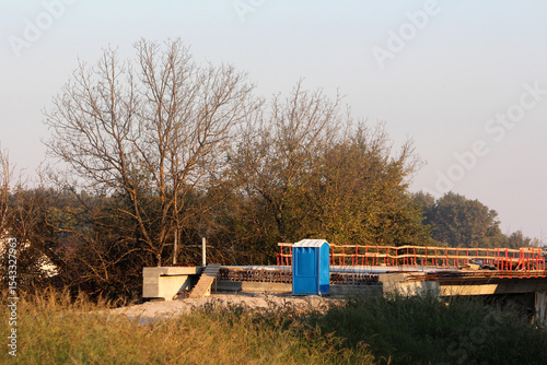 Bright blue portable toilet stands on a concrete slab at a rural construction site, surrounded by tall grass, bare autumn trees, and orange safety railings in the evening light
