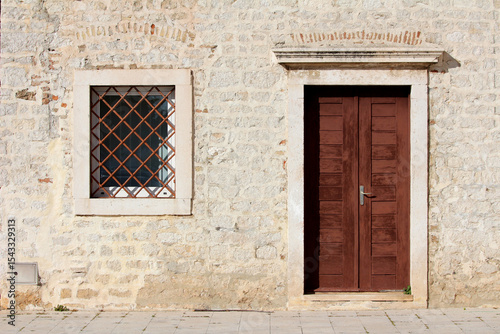 Historic stone building features a double wooden door and a square window with metal bars, framed by light stonework and illuminated by direct sunlight on a paved street