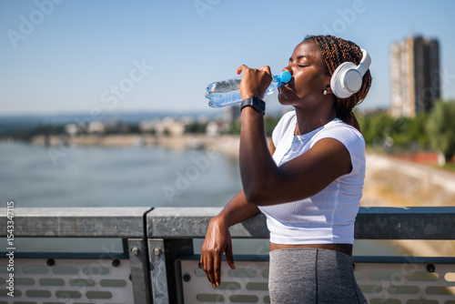 Portrait of fit sporty African-American woman with  headphones in sports clothing drinking water while standing at the bridge during sport training.