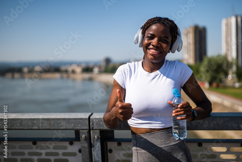 Portrait of  young sporty African woman in sportswear with headphones drinking water and showing thumb up while taking a break from exercising on the city bridge.