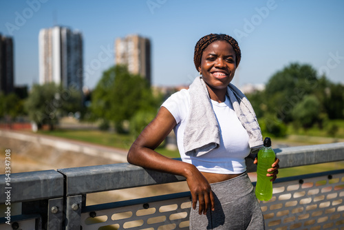 Portrait of young sporty African-American woman in sports clothing drinking energy drink while resting after exercising outdoor on a sunny day.