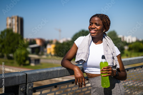 Portrait of young sporty African woman in sportswear drinking energy drink while enjoying taking a break from exercising on the city bridge.