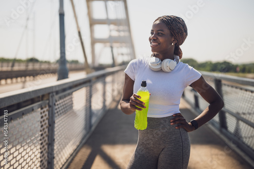 Portrait of young sporty African-American woman in sport clothing with headphones drinking energy drink after sport training on urban city bridge.