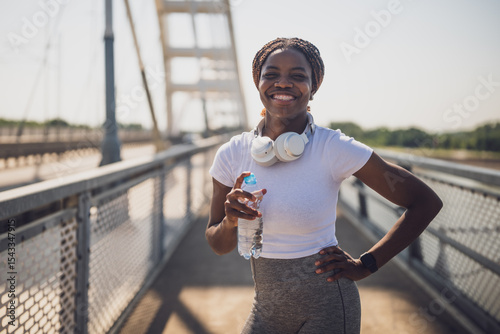 Portrait of sporty African-American woman in sportswear with headphones drinking water during exercising on urban city bridge.