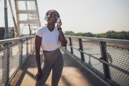 Joyful African-American sporty woman in sports clothing enjoying listening to music through headphones during sport training on urban city bridge.