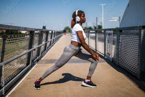 Motivated young African-American woman in sportswear with headphones enjoying exercising and stretching while standing at the city bridge. She is in the squatting position.