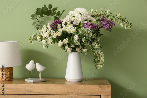 Vase of beautiful flowers bouquet with lamp on commode near green wall in stylish spring bedroom
