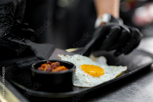 Wallpaper Mural Close-up of a gloved hand serving a sunny-side-up egg with pepper beside cooked beans on a black plate, captured in a dark, moody culinary setting. Torontodigital.ca