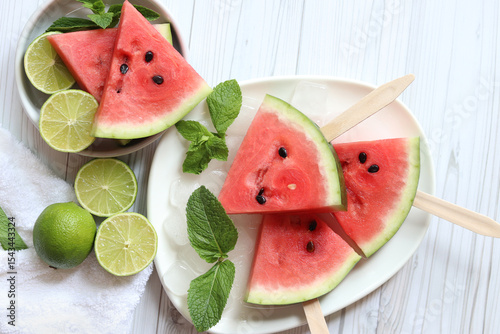 Ripe watermelon pieces on sticks like ice cream. Watermelon, lime and mint. Summer fruits background. Flat lay. Watermelon slices in white plate top view, wooden table and lime halves. Summer food