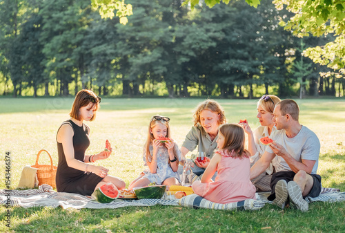 Big family sitting on the picnic blanket in city park during weekend Sunday sunny day. They are smiling, laughing and eating boiled corn and watermelon. Family values and outdoors activities concept.