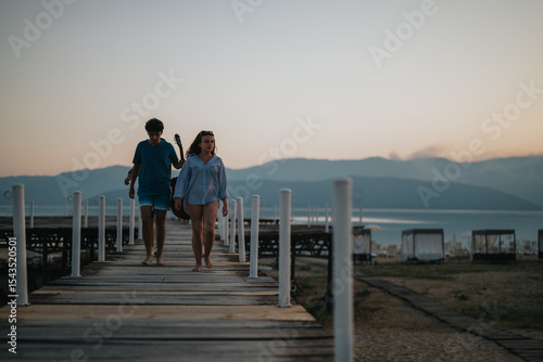 A pair of friends stroll along a wooden pier, with scenic mountains and calm water providing a tranquil backdrop during sunset.