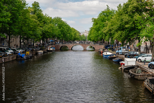 Amsterdam, Netherlands – May 21, 2025: A long view of an Amsterdam canal, framed by lush trees and residential buildings, with a prominent arch bridge in the middle ground