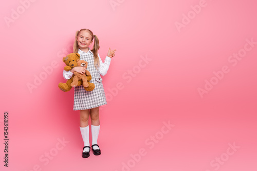 Smiling schoolgirl wearing a stylish uniform holding a teddy bear on a pink background showing a peace sign photo