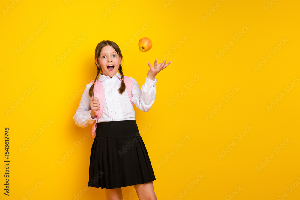 Adorable schoolgirl with backpack bouncing an apple in delight while dressed in school uniform on vibrant yellow background Adorable schoolgirl with backpack bouncing an apple in delight while dressed in school uniform on vibrant yellow background