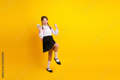 Cheerful schoolgirl holding a phone and posing on a yellow background in stylish uniform with a backpack