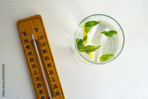 Glass of detox water with ice and citrus near thermometer. Healthy lifestyle: hydration in summer heat. Body cooling methods, temperature stress and refreshing natural beverages for wellness.