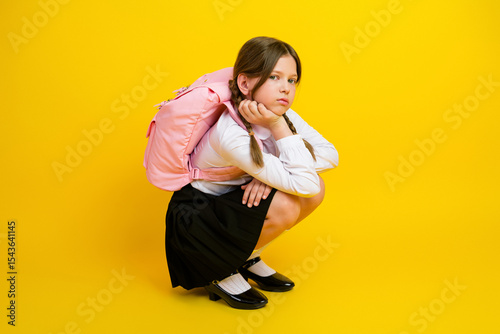 Schoolgirl sitting against bright yellow background wearing a school uniform and pink backpack, looking thoughtful and stylish